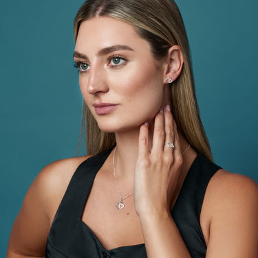Woman wearing heart shaped jewellery including earrings, a necklace, and a ring against a teal background