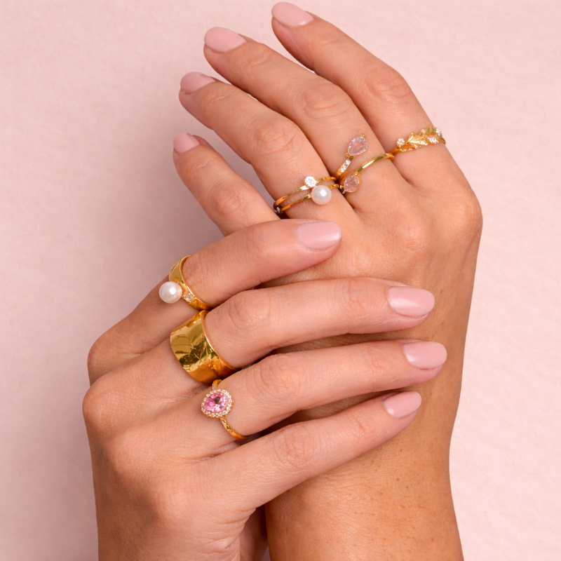 Hand wearing multiple gold rings with gemstones on a pink background